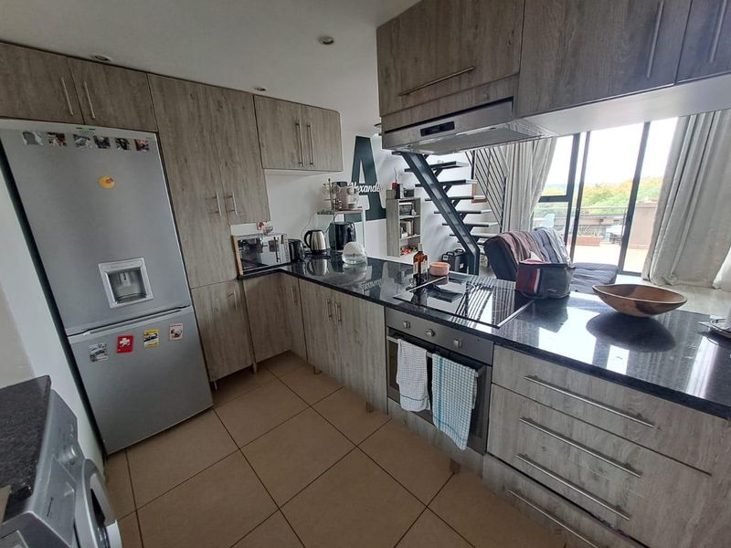 Modern kitchen featuring wood-look cabinetry, granite countertops, integrated appliances, and tiled flooring, seamlessly flowing into the living area with staircase access.