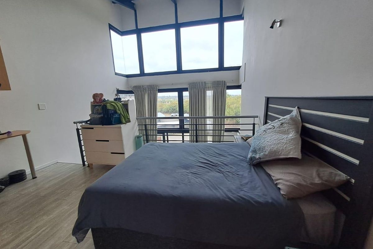 Spacious loft-style bedroom featuring high ceilings with exposed beams, large windows providing ample natural light, and a mezzanine-style metal balustrade overlooking the lower level.