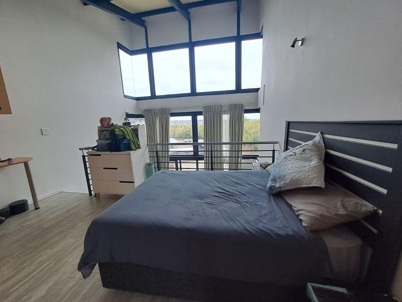 Spacious loft-style bedroom featuring high ceilings with exposed beams, large windows providing ample natural light, and a mezzanine-style metal balustrade overlooking the lower level.