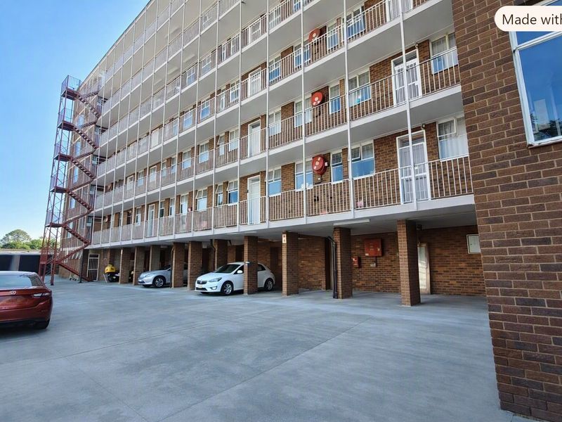 Exterior view of a multi-story apartment building featuring brick facade, private balconies with metal railings, and a ground-level covered parking area.