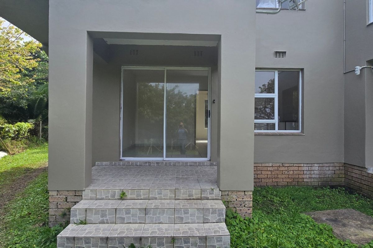 Rear exterior view of the property featuring a tiled patio entrance, sliding glass doors, and brick base detailing set against a garden area.