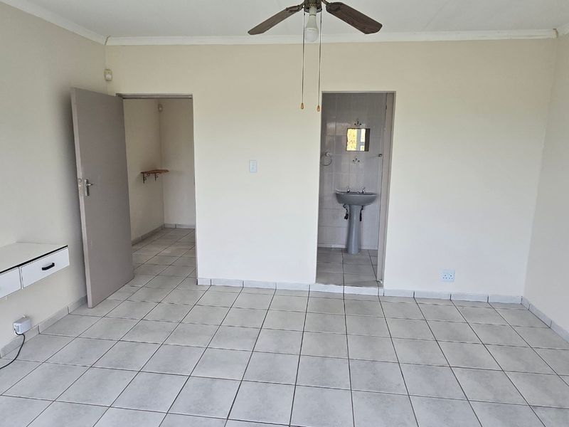 A spacious, empty bedroom featuring tiled flooring, a ceiling fan, a built-in wall desk, and an en-suite area with a pedestal basin.