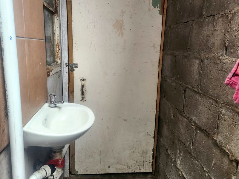A small utility area featuring a wall-mounted basin, exposed plumbing, and unfinished concrete block walls, requiring significant maintenance and cosmetic updates.