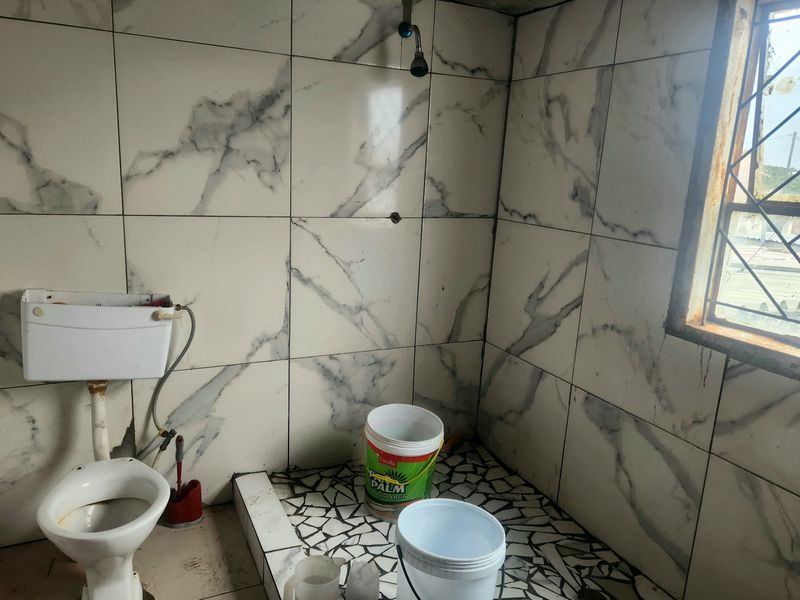 A small bathroom featuring marble-effect wall tiles, mosaic flooring, a basic toilet, and a wall-mounted shower head, requiring significant renovation and maintenance.