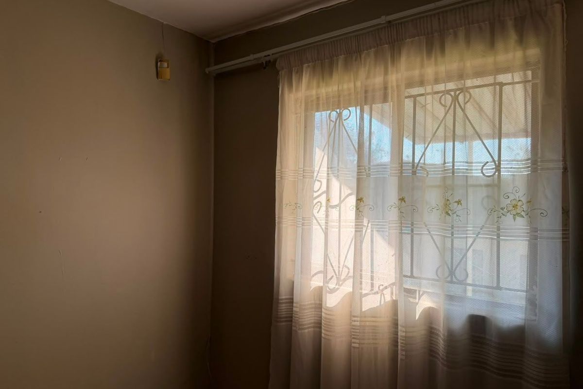 A simple bedroom featuring a window with a security gate and an installed alarm sensor, providing a functional and secure space.