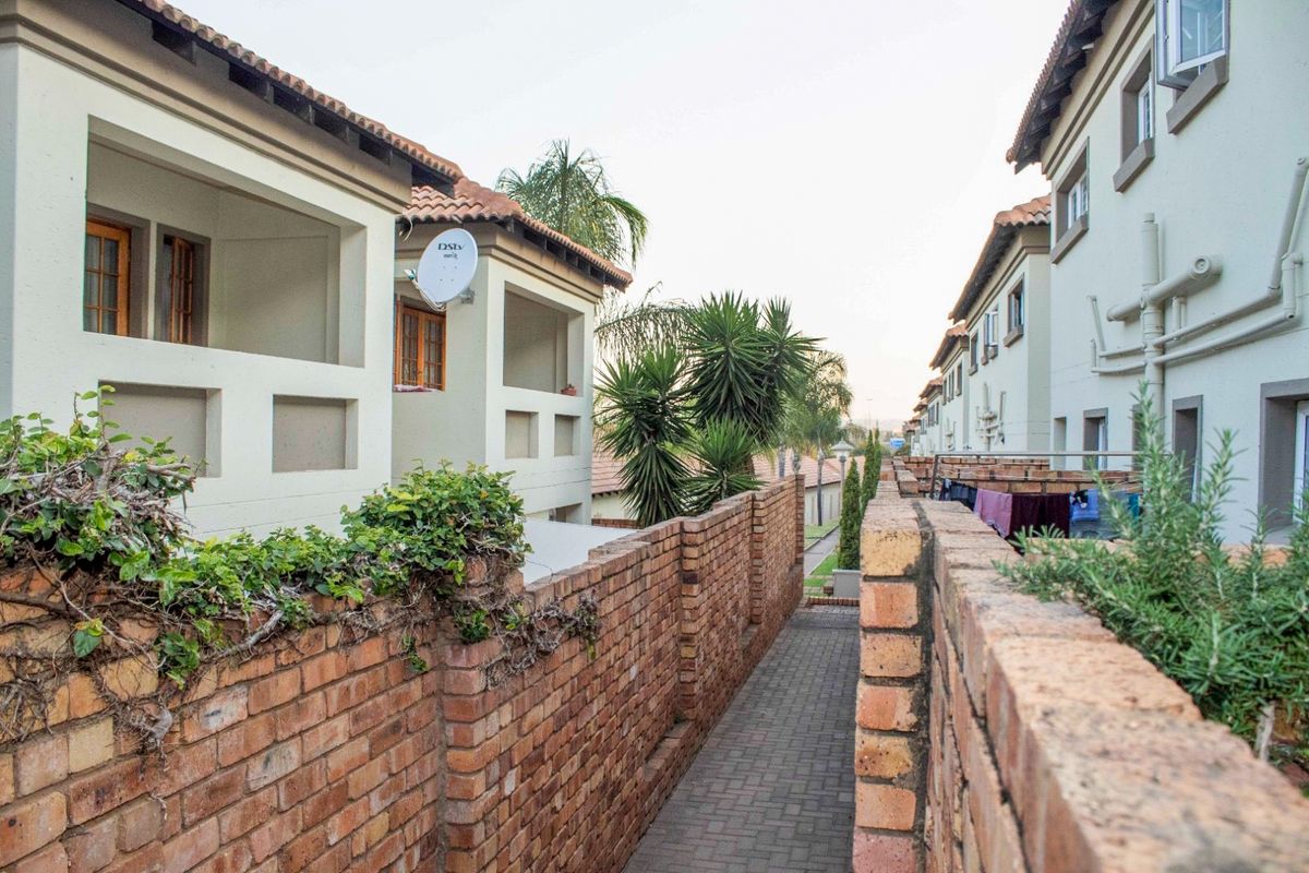 A view of the exterior rear walkway between townhouse units, featuring brick boundary walls, paved pathways, and upper-level balconies.