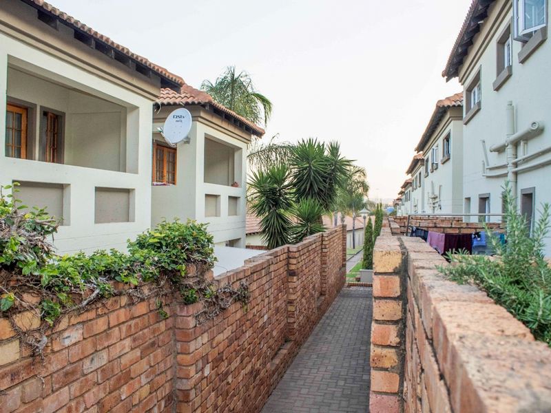 A view of the exterior rear walkway between townhouse units, featuring brick boundary walls, paved pathways, and upper-level balconies.