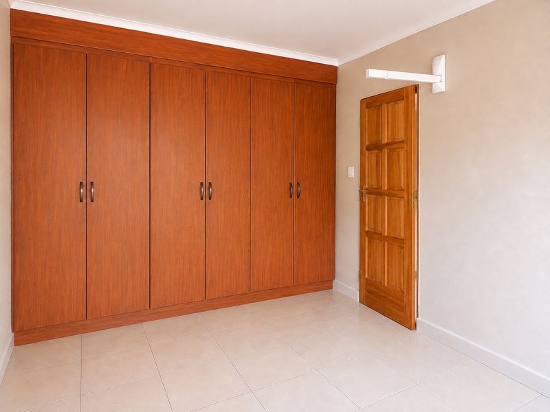 A clean, empty bedroom featuring extensive built-in wooden cupboards, neutral tiled flooring, and a solid wooden door.