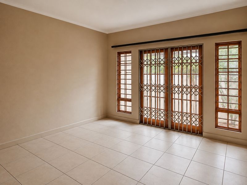 A bright, empty living room featuring neutral tiled flooring, wooden window frames, and sliding glass doors secured with metal security gates.