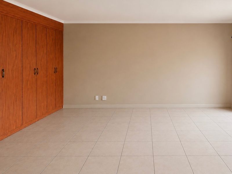 An empty bedroom featuring neutral tiled flooring and extensive built-in wooden cupboards, providing a clean and versatile space ready for personalization.