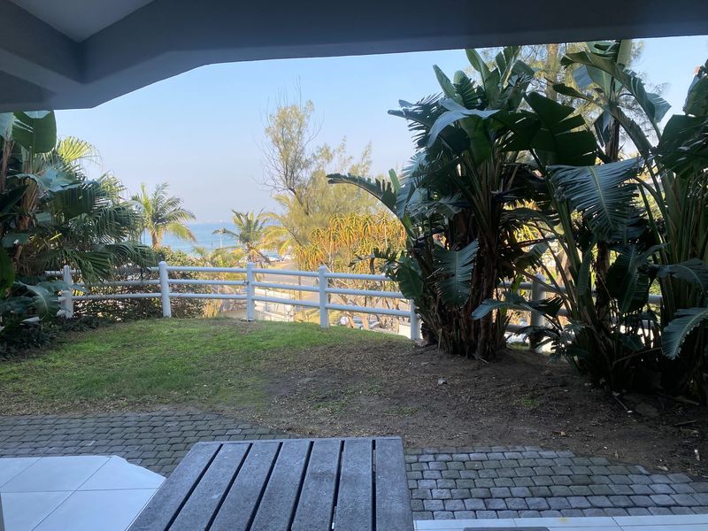 A covered patio area featuring paved flooring and a white perimeter fence, overlooking a private garden with established tropical plants and distant sea views.