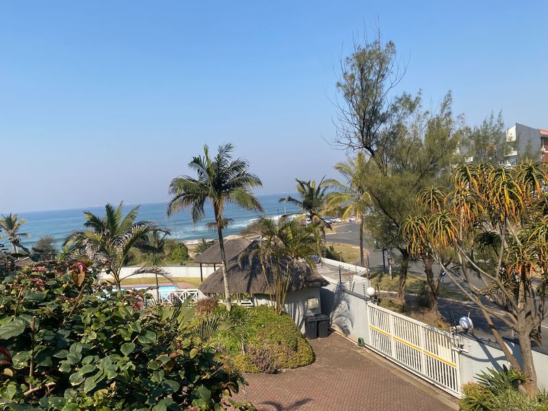 An elevated view overlooking the property grounds, featuring a paved driveway, secure gate, thatched structure, and a scenic coastal outlook.