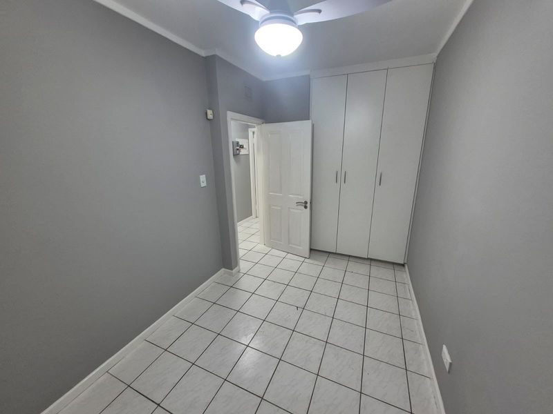 A clean, empty bedroom featuring neutral grey walls, white tiled flooring, and built-in white cupboards with a ceiling fan.