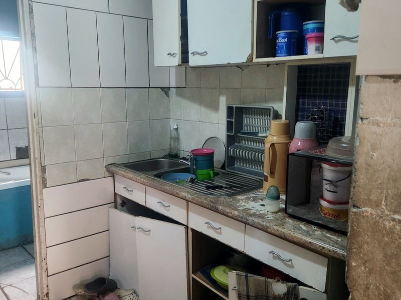 A dated kitchen area featuring built-in cabinetry, a stainless steel sink, and tiled splashbacks, currently showing significant signs of wear and unfinished structural elements.