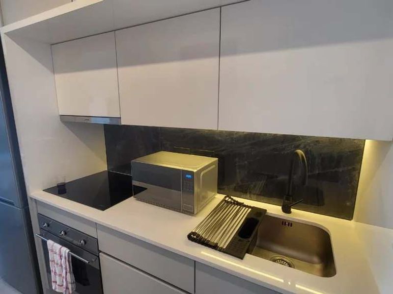 Modern kitchen featuring sleek white cabinetry, stone countertops, dark stone splashback, integrated hob, oven, and high-quality black fixtures with under-cabinet lighting.