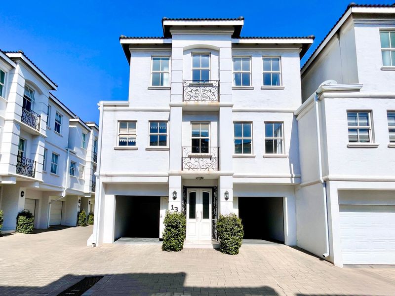 A well-maintained three-story townhouse featuring a double garage, paved driveway, and decorative wrought iron Juliet balconies on the upper levels.
