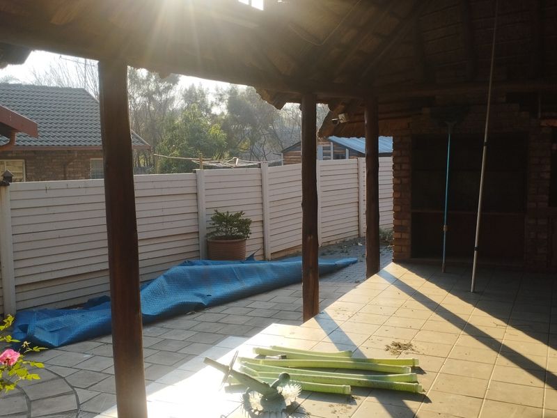 A covered outdoor patio area featuring a thatched roof, timber support posts, and a built-in brick braai, overlooking the rear garden.