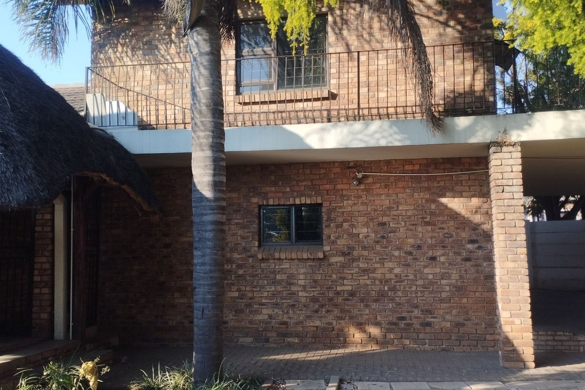Rear view of a double-story face brick house featuring a balcony with metal railings, a thatched roof section, and a paved patio area.