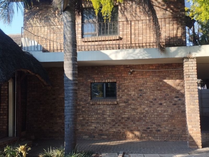 Rear view of a double-story face brick house featuring a balcony with metal railings, a thatched roof section, and a paved patio area.