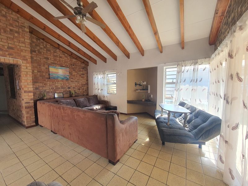 Spacious living room featuring exposed timber ceiling beams, a rustic brick feature wall, tiled flooring, and natural light through louver windows.