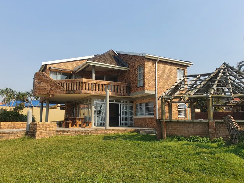 Rear view of a double-story face brick house featuring a balcony, ground floor patio, and a weathered thatched lapa structure in the garden.