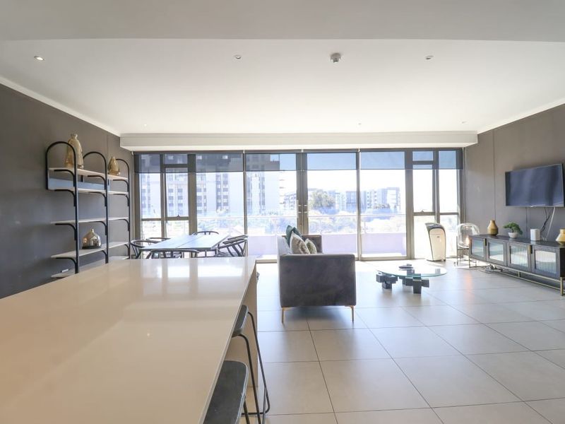 Spacious open-plan living and dining area featuring large format floor tiles, a modern kitchen island, and expansive floor-to-ceiling glass doors leading to a balcony.