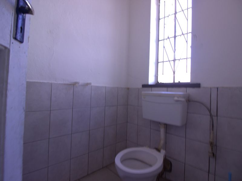 A compact bathroom featuring a standard toilet, tiled walls, and a window with security bars, requiring cosmetic updates to improve its overall condition.