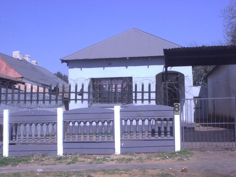 Front elevation of a single-story house featuring a corrugated metal roof, security fencing with razor wire, and a gated carport entrance.