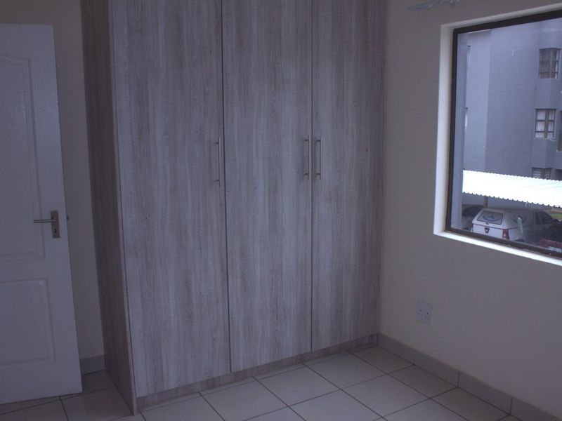 A clean, empty bedroom featuring light wood-finish built-in wardrobes, neutral tiled flooring, and a window providing natural light.