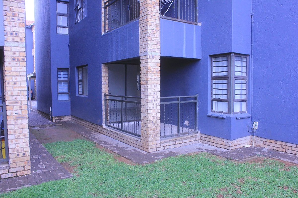 Rear view of the apartment building featuring a ground floor patio with metal railings, brick pillars, and surrounding paved areas with lawn.