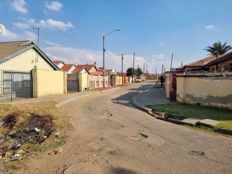 A street view showing the residential neighborhood approach with paved roads, perimeter walls, and street lighting.