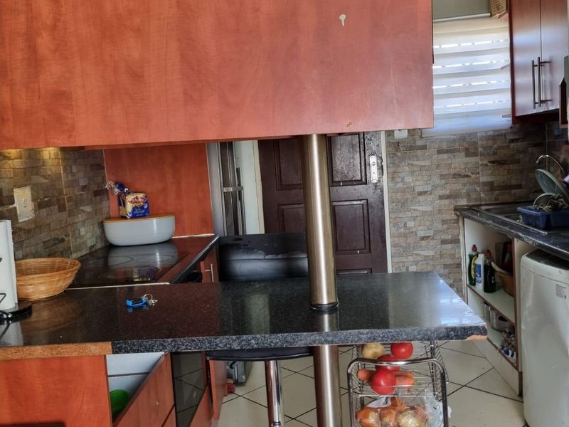 Compact kitchen featuring laminate cabinetry, a breakfast bar with a stainless steel support column, stone-look backsplash, and ceramic floor tiles.