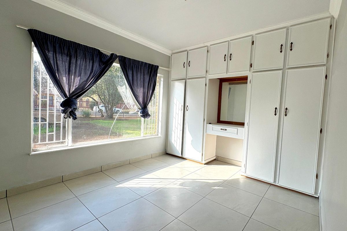 A bright, empty bedroom featuring extensive built-in white cabinetry with an integrated vanity, tiled flooring, and large windows with security bars.