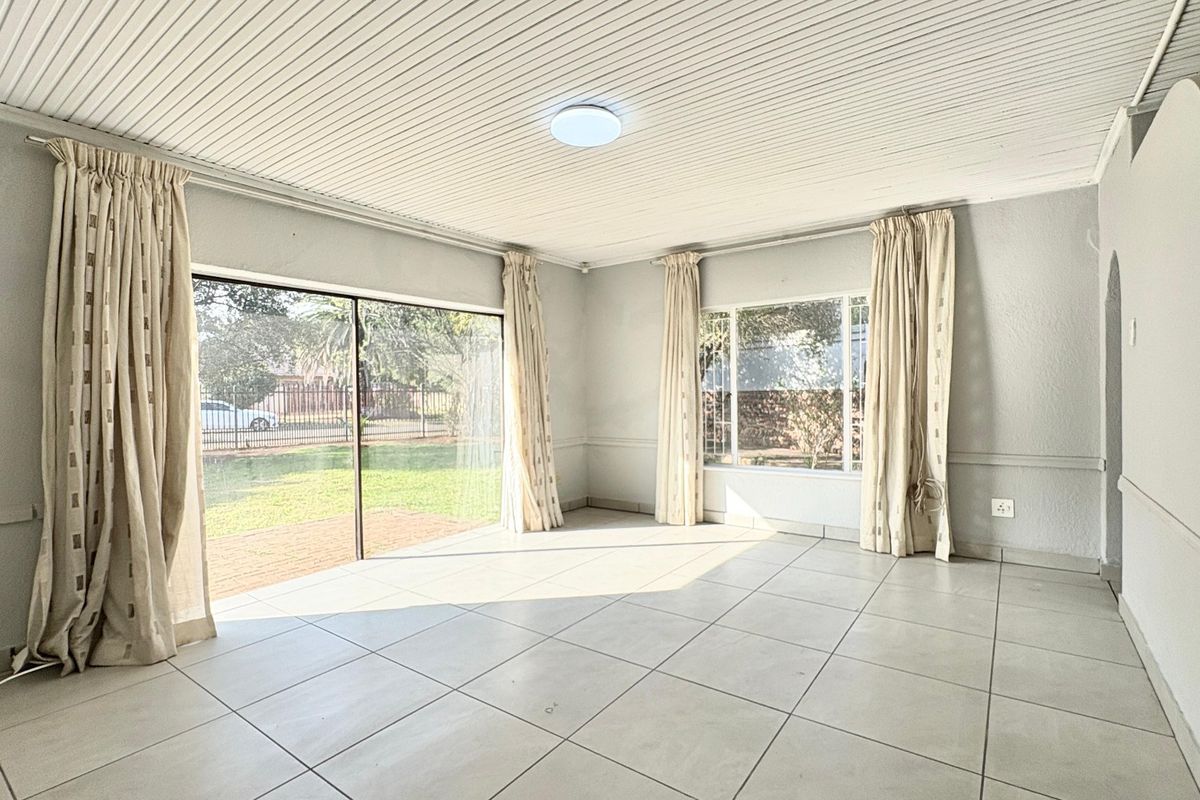 A bright, empty living room featuring large windows, sliding glass doors to the garden, tiled flooring, and a classic tongue and groove ceiling.