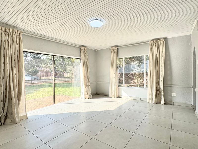 A bright, empty living room featuring large windows, sliding glass doors to the garden, tiled flooring, and a classic tongue and groove ceiling.