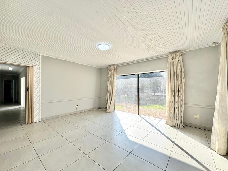 A bright, empty living room featuring large sliding glass doors, neutral tiled flooring, and a classic tongue and groove ceiling.