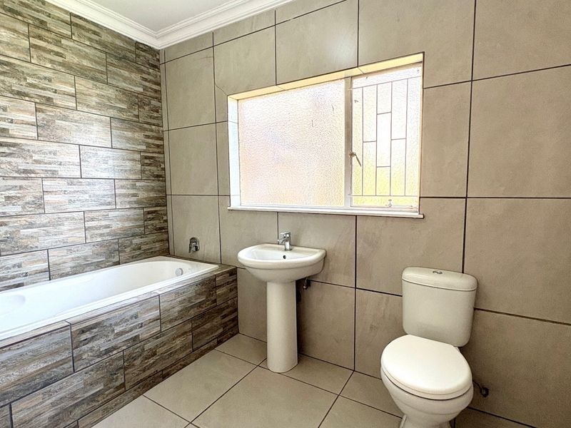 A clean, modern bathroom featuring a built-in bathtub, pedestal basin, toilet, and neutral floor-to-ceiling tiling.