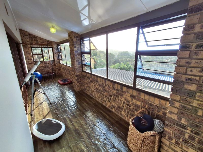 A sunlit enclosed balcony featuring exposed brick walls, tiled flooring, and expansive windows offering an elevated view of the surrounding greenery.