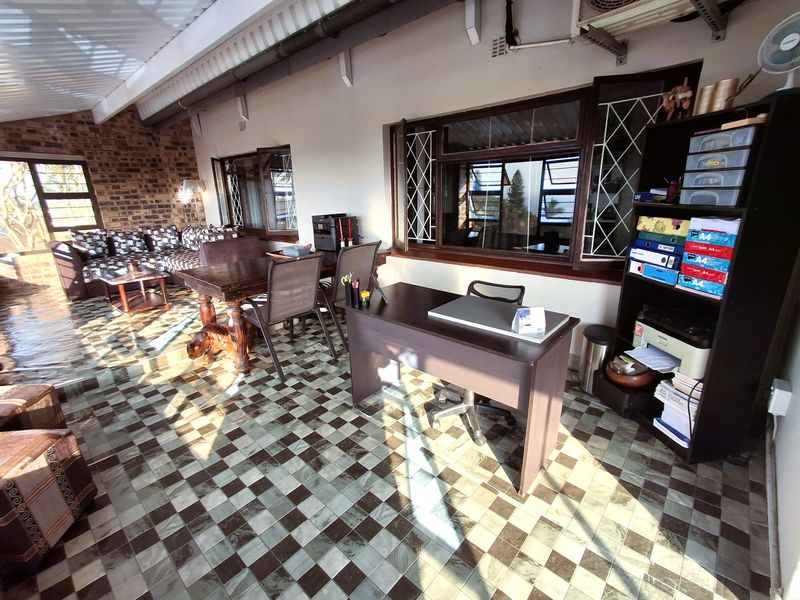 A spacious covered patio area featuring exposed brickwork, tiled flooring, and large windows with security bars, suitable for a multi-purpose living or workspace.