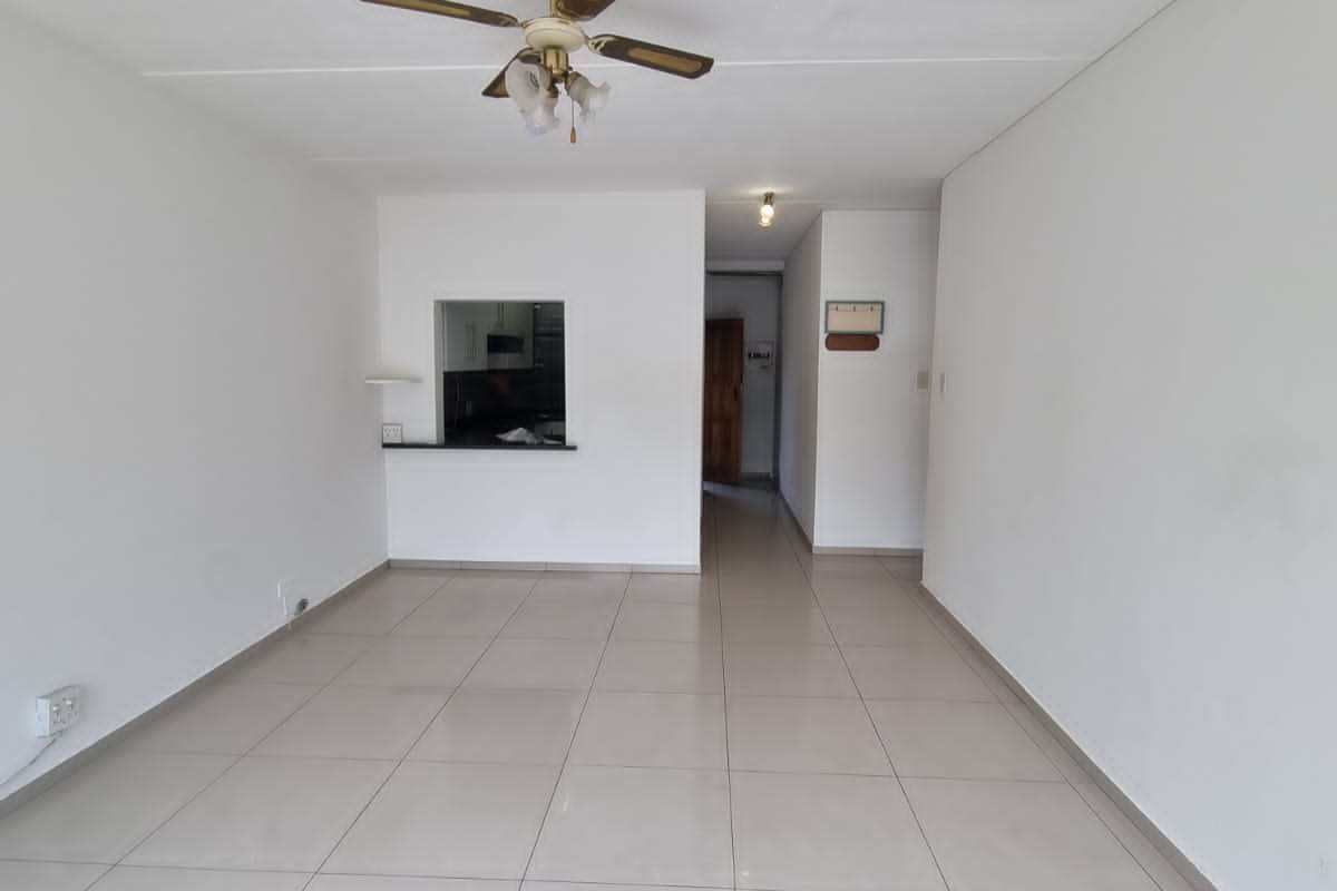 An empty, open-plan living area featuring neutral tiled flooring, a ceiling fan, and a convenient pass-through window connecting to the kitchen.