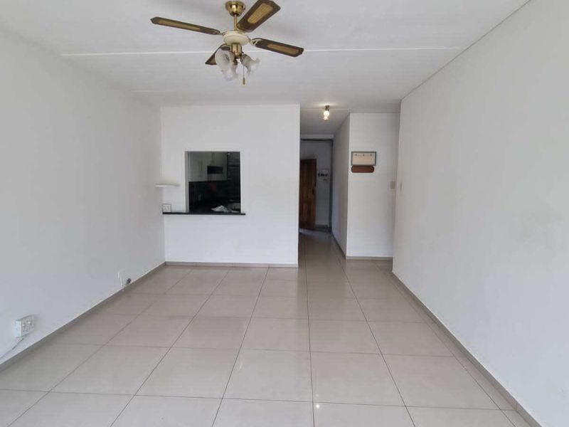 An empty, open-plan living area featuring neutral tiled flooring, a ceiling fan, and a convenient pass-through window connecting to the kitchen.