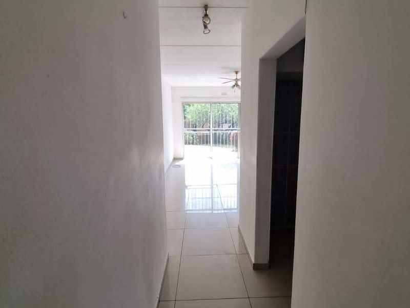 A vacant hallway with tiled flooring leading towards a bright living area with a sliding glass door and ceiling fan.