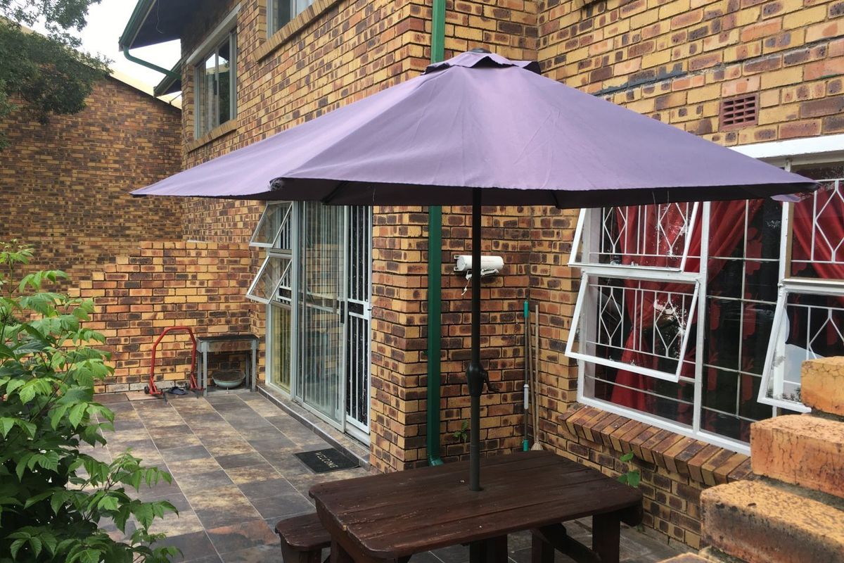 A rear exterior view of a face brick townhouse featuring a tiled patio area, security-fitted windows, and a sliding door entrance.