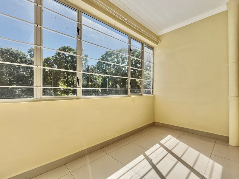 A bright, empty enclosed balcony or sunroom featuring large windows with security bars, tiled flooring, and neutral wall finishes.