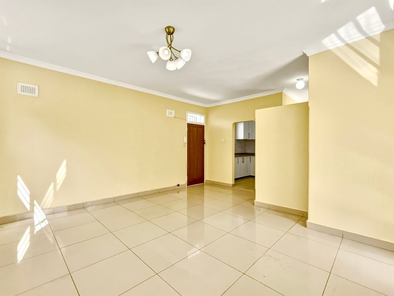 A bright, empty living area featuring neutral tiled flooring, coved cornicing, and a direct view into the adjacent kitchen space.