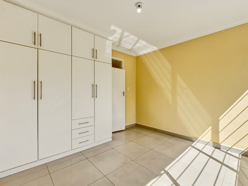 A bright, empty bedroom featuring neutral yellow walls, large built-in white cupboards, and clean tiled flooring with natural light.