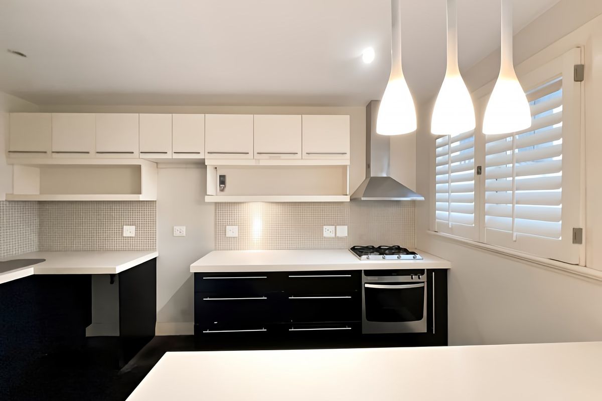A modern kitchen featuring black lower cabinetry, white upper cabinets, mosaic tile backsplash, built-in gas cooktop, oven, and elegant pendant lighting.