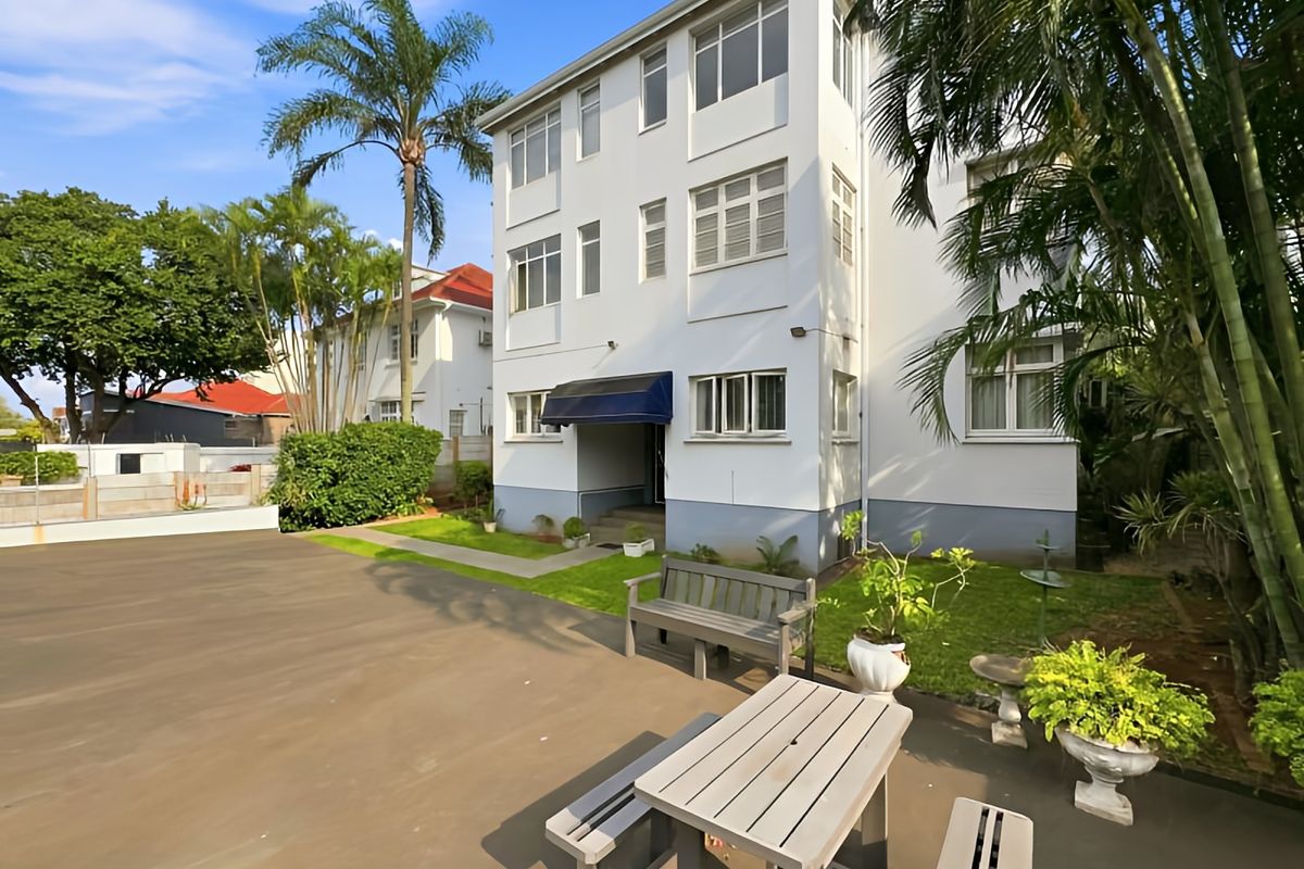 A view of the rear elevation of a multi-story apartment building featuring a paved courtyard area and established garden surroundings.