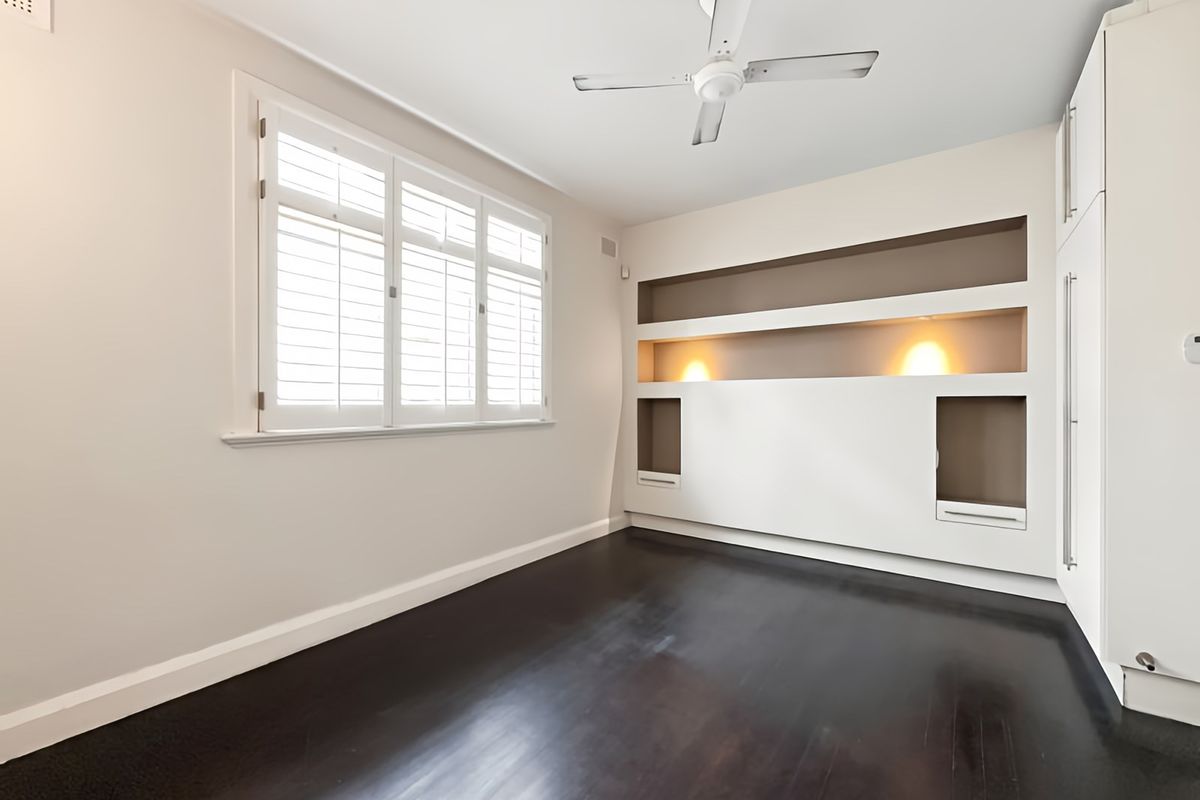A bright, empty bedroom featuring dark timber flooring, plantation shutters, a ceiling fan, and custom built-in shelving with integrated accent lighting.