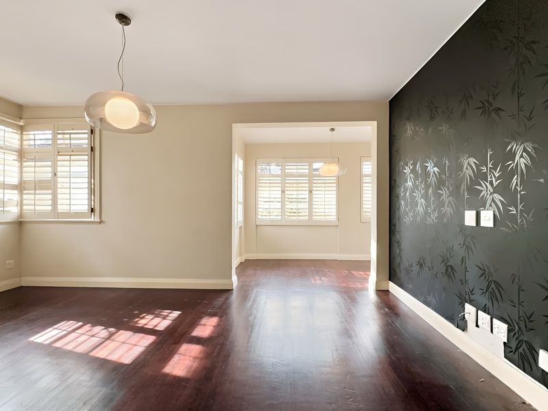 Spacious, light-filled living area featuring polished hardwood floors, plantation shutters, and a striking feature wall, offering a versatile and well-maintained open-plan layout.